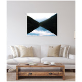 Framed photograph above sofa showing mountain slopes mirrored symmetrically in a still lake at sunrise, with soft light peeking over dark peaks.