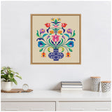 Framed canvas print featuring a symmetrical floral arrangement with colorful stylized flowers in a patterned blue vase on a beige background, hanging above a white shelf with plants and books.