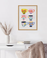 Framed print featuring six colorful teacups with distinct botanical patterns, displayed above a white desk with a vase of pampas grass and stacked books.