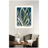 Abstract botanical print with layered green and beige leaves on a deep blue background, framed in light wood and hung above a brown round wooden table and dark armchairs.