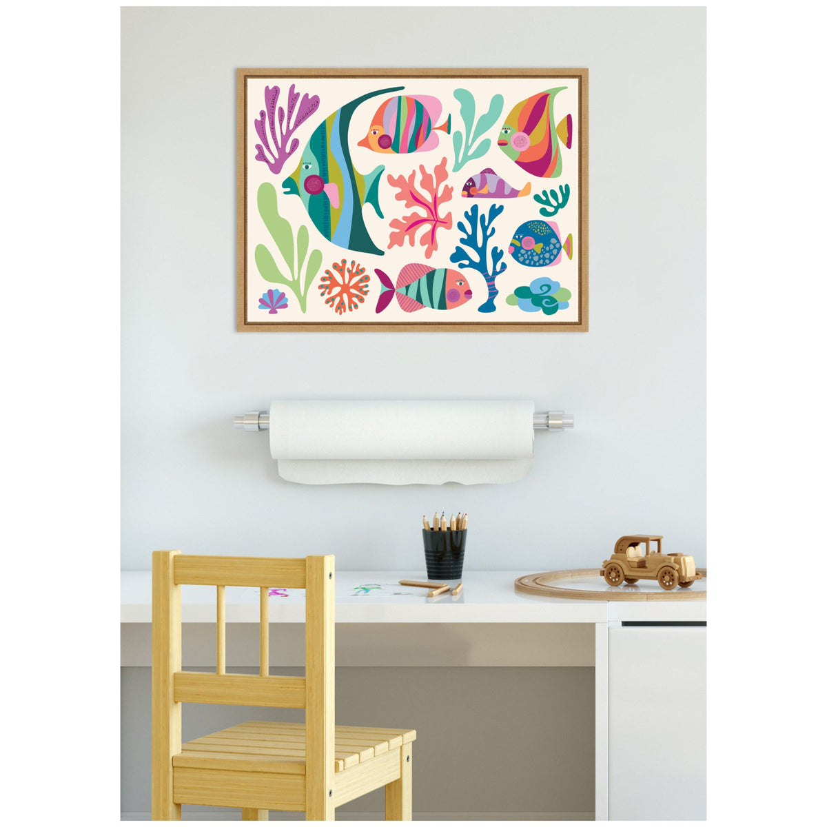Framed canvas with colorful tropical fish and coral illustrations in pink, blue, green, and orange, hanging above a white desk with wooden chair and toys.