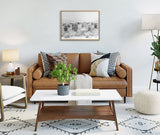 Black and white framed photo of a herd of Highland cows with tall grass, mounted above a brown leather sofa adorned with patterned pillows in a bright living room.