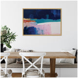 Abstract painting with layered blocks of dark blue, turquoise, pink, and touches of gold, framed in light wood, hanging above a wooden dining table with white chairs.