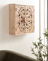 Light wood wall-mounted cabinet with intricate round floral carving centered on double doors, shown angled with green couch and leafy plant nearby.
