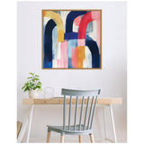 Abstract framed wall art with bold arches in navy blue, red, mustard, and pink hues above a minimalist wooden desk and a pale gray chair.