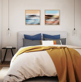 Modern bedroom with a beige upholstered headboard, blue and patterned pillows, yellow knitted throw on cream bedding, and two framed abstract landscape prints above.