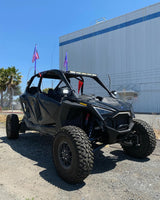 Black Polaris Pro R RZR off-road vehicle with large knobby tires, roof-mounted light bar, and two flags attached to rear, parked on gravel beside industrial building.