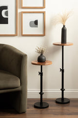 Two adjustable plant stands with warm wood tops and matte black metal bases, styled with a green velvet armchair and monochrome framed art on a white wall.