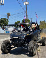 Black Polaris Pro R RZR 4-seater off-road vehicle with large knobby tires and a light bar mounted on the roof, parked beside a road under a clear blue sky.