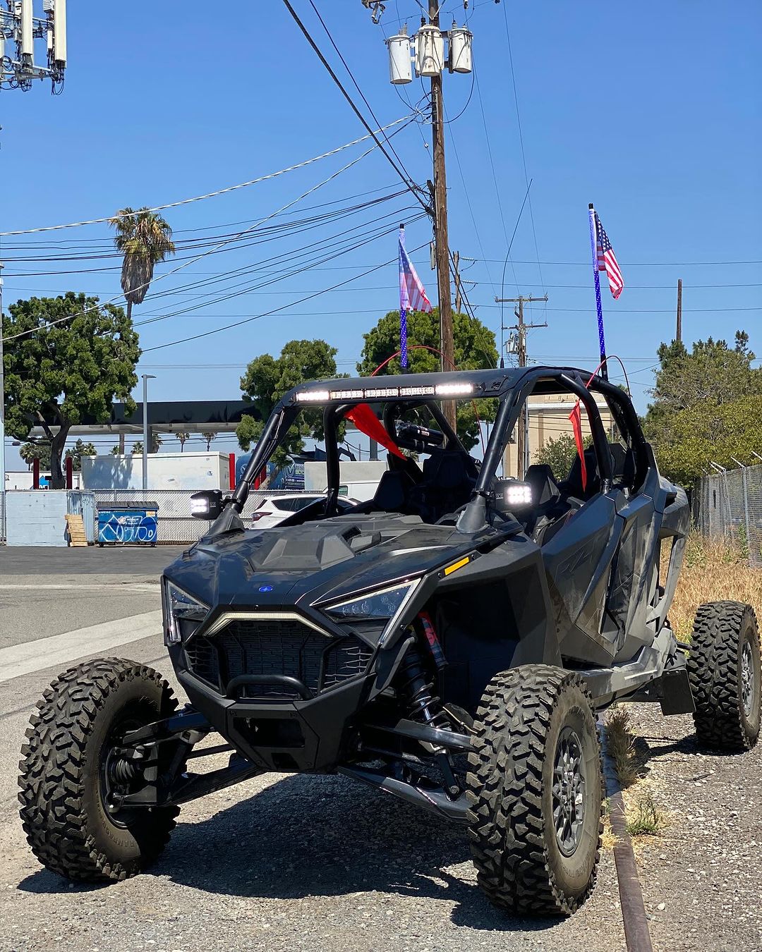 Black Polaris Pro R RZR 4-seater off-road vehicle with large knobby tires and a light bar mounted on the roof, parked beside a road under a clear blue sky.