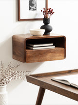 Wall-mounted wooden floating side table with rectangular curved edges, one drawer, and open shelf holding two books, complemented by black vase with red buds and white bowl on top.