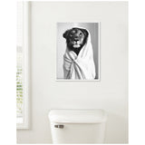 Black and white canvas of a lioness wrapped in a white towel hanging above a white toilet against a plain wall.