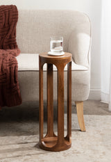 Tall, cylindrical wooden drink table with vertical cut-out panels, placed beside a beige sofa with a brown knitted throw, holding a glass of water on a coaster.
