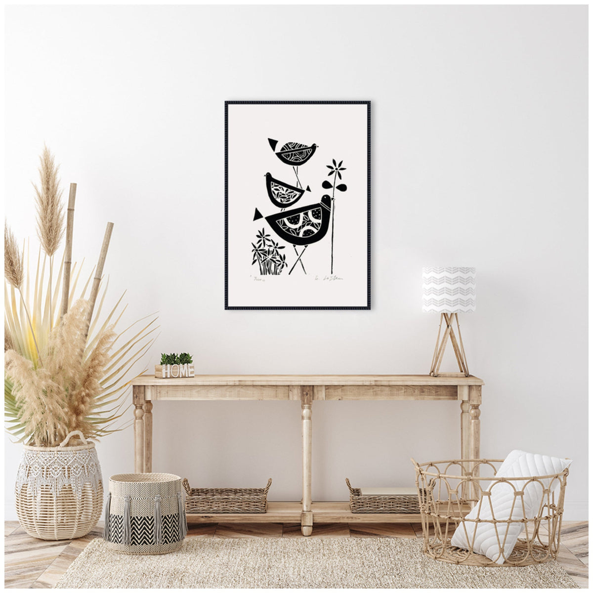 Black and white linocut artwork of three stylized birds stacked with foliage, framed on a white wall above a rustic wooden console in a minimalist room.