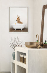 Framed canvas of a brown baby donkey sitting in a white clawfoot bathtub filled with bubbles, hanging above a minimalist bathroom counter with a gold sink and faucet.