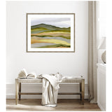 Framed landscape artwork with layers of green, yellow, and muted purple hills hanging above a wooden bench with a white open book and draped cream fabric in a bright, minimalist room.