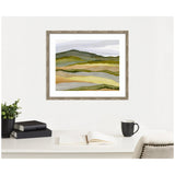Framed abstract landscape painting showing layered hills in muted green, yellow, and gray tones, hanging above a white desk with books, a plant, an open notebook, and a black mug.