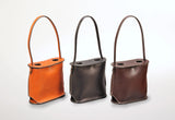 Three leather wine totes in orange, dark brown, and medium brown, each with two bottle holes and a rounded handle extending from the rectangular base.