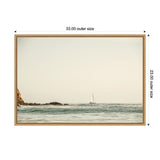 Sailboat on calm ocean near rocky coastline under a pale sky, framed in light wood with outer size dimensions 33 by 23 inches.