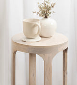 Round light wood side table with smooth edges, holding a cream ceramic cup on a matching saucer and a small white vase with dried flowers, set against a white curtain.