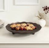 Dark brown decorative tray with scalloped edges and three short legs holding croissants and cranberries on a white surface, flanked by white vases with dried flowers.