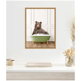 Framed canvas art showing a brown bear sitting in a vintage green bathtub filled with white bubbles against a white paneled wall and wooden floor.