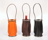 Three cylindrical leather wine totes in orange, dark brown, and black with long looped handles and visible stitching, one holding a red wine bottle.