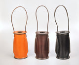 Three leather wine totes in orange, dark brown, and black, each with a cylindrical shape, top carrying loop, and visible contrasting stitching.