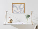 White wall-mounted shelf with leather strap supports holds a gold geometric vase with brushes, a small gold dish, an open notebook, and a vase with green leaves, below framed abstract wave print.