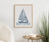 Framed blue sailboat print with textured sails above stylized water, hung on a white wall next to a round brass side table and green plant.
