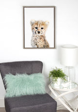 Framed canvas portrait of a young cheetah with soft fur and dark tear marks, hanging above a gray chair with a mint green fluffy pillow and a white side table with a clear lamp and small green plant.