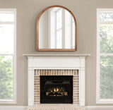Arched wood-framed wall mirror with warm natural finish reflecting a bright window, hung above a white mantel and brick fireplace with a fire burning inside.