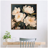 Square framed floral painting of four large creamy white peonies with green leaves on a dark background, hung above a wooden desk with a plant and metal sculpture.
