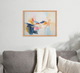 Abstract coastal-inspired painting with soft pinks, blues, yellows, and white, framed in wood, hanging above a gray sofa with a white textured throw and pillow.