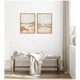 Two framed canvas paintings of soft coastal landscapes with sandy dunes and blue-green water, hung side by side on a white wall above a rustic wooden bench with an open book and draped fabric.