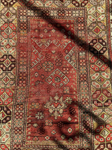 Antique Turkish wool prayer rug featuring a central faded red field with geometric and floral motifs, bordered by intricate hexagonal and diamond patterns in red, cream, and brown.