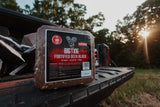 Rectangular block of Big Tine Fortified Deer Block bait with a red and black label, placed on the tailgate of a pickup truck outdoors during sunset.