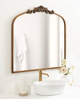 Traditional arched mirror with a carved floral crest and warm wood frame above a white oval vessel sink and gold faucet, set against a light wall with white towel background.