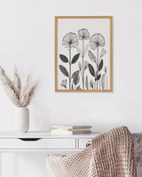 Black and white linocut print of stylized flowers with intricate line patterns in a light wooden frame against a white wall above a white desk with books and a white vase with pampas grass.