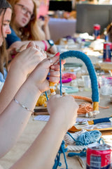 Close-up of hands wrapping blue yarn around a circular frame during a crafting activity with colorful materials and drinks on the table in soft focus background.