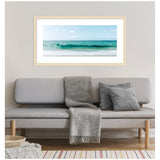 Framed horizontal photograph of a turquoise ocean wave under a blue sky with scattered clouds, hanging above a minimalist gray sofa with a gray throw and cushions.