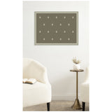 Green botanical block print with white floral motifs arranged in a grid, framed and hung on white wall above a pair of cream-colored armchairs and brass side table with white vase.