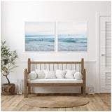 Two framed canvases above a wooden bench with white cushions, depicting calm ocean scenes under soft, cloudy skies with birds in flight.