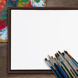 Blank square canvas with a dark wooden frame resting on a wooden surface, with a cluster of paintbrushes and a partially visible colorful paint palette nearby.