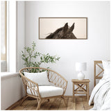 Framed canvas print featuring a close-up of a black horse's ears and mane against a soft beige background, hung above a wicker chair and wooden side table with white lamp.
