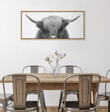 Framed black-and-white close-up of a Highland cow's face with long horns and shaggy hair above a wooden dining table with metal chairs and a glass vase with dried stems.