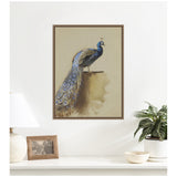 Framed canvas print of a peacock with dark blue and gold feathers perched against a beige background hanging above a white tabletop with a lamp, photo frame, and potted plant.