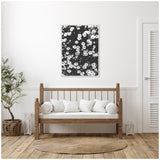 Black and white floral canvas artwork hanging above a wooden bench with white cushions, flanked by a woven basket planter and a lantern on light wood flooring.