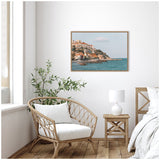 Coastal town with terracotta-roofed buildings on a rocky hillside beside blue sea, framed and hung on a white wall above a rattan chair and wooden side table.