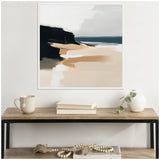 Framed abstract coastal landscape painting with dark cliffs, sandy beige shore, and muted blue-gray ocean and sky above a wooden console table with a mug, books, and a white vase with green leaves.
